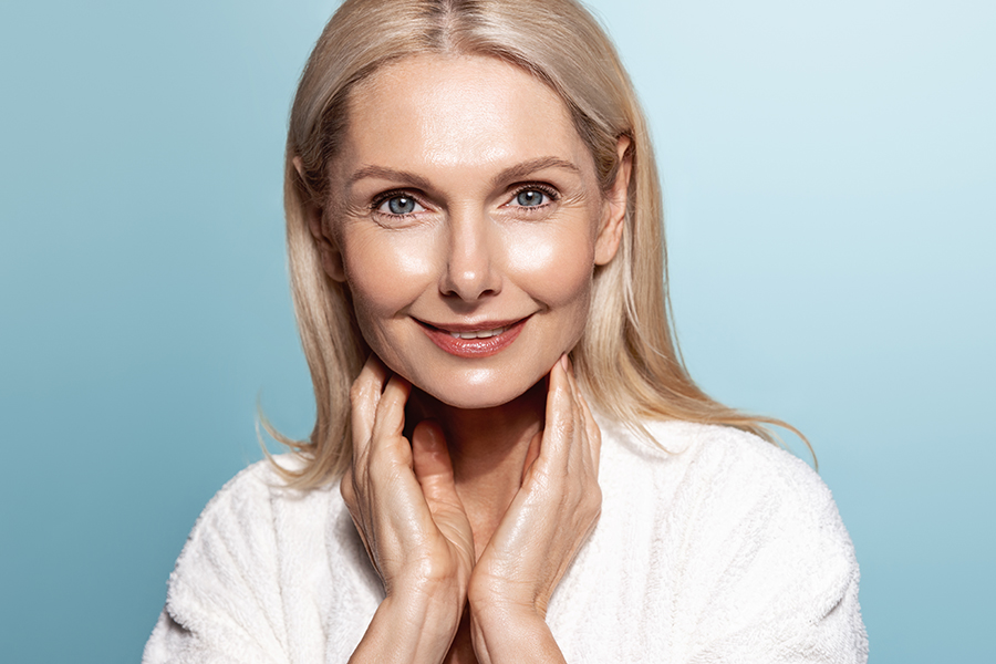 Smiling s middle aged mature older woman applying facial cream on face looking at camera isolated on blue background. Anti age healthy dry skin care beauty therapy concept, old skincare treatment
